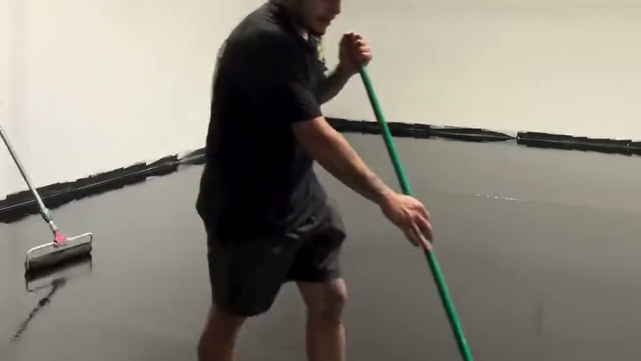An installer rolling a wet epoxy base coat across a large commercial shop floor, the wall reflected in the surface behind him.