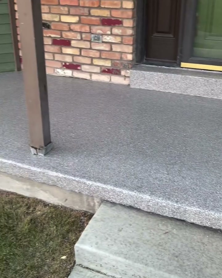 A finished front porch coated in a light grey pumice flake blend, meeting brickwork and a lawn edge.