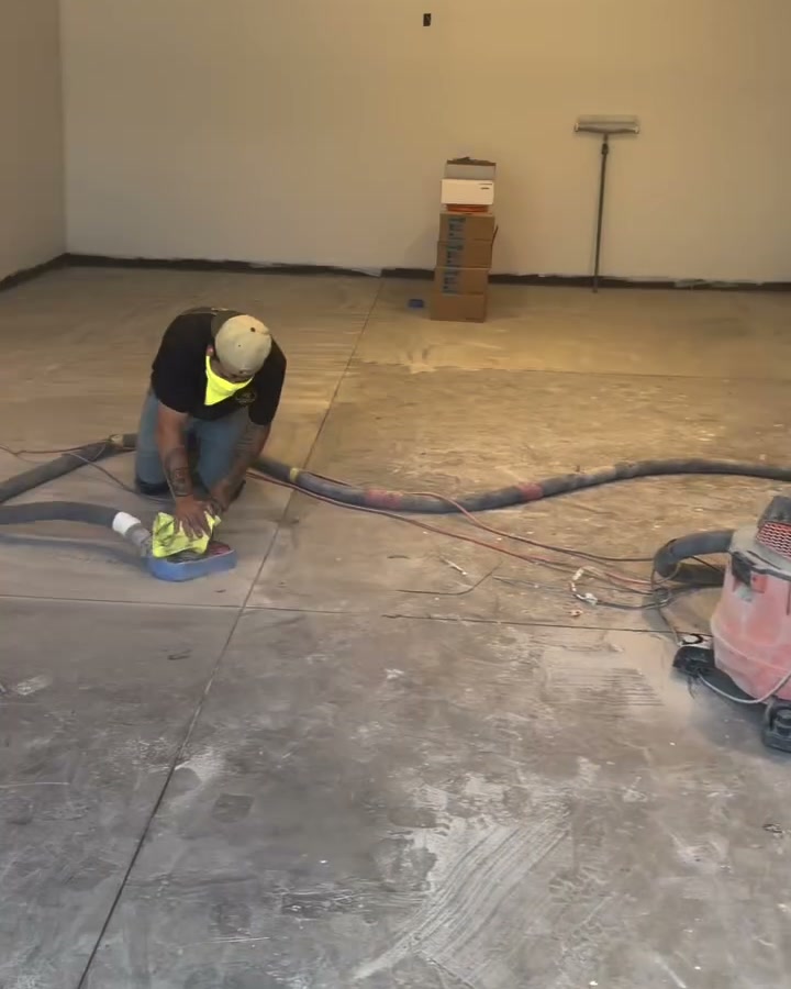 An installer kneeling to grind a bare concrete shop slab with a hand grinder and a vacuum hose behind him.